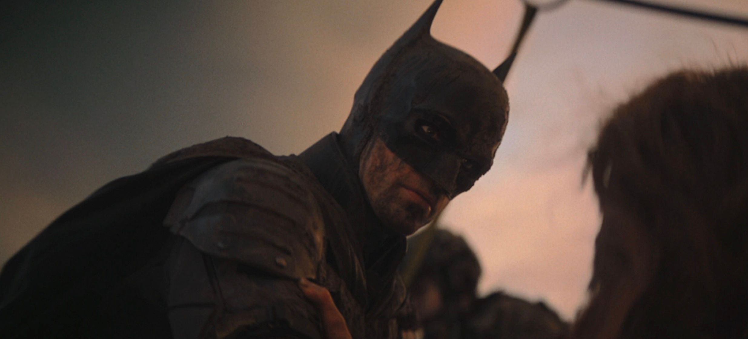 A very grubby-looking Batman (Robert Pattinson) looks down at a prone woman he’s rescuing in The Batman