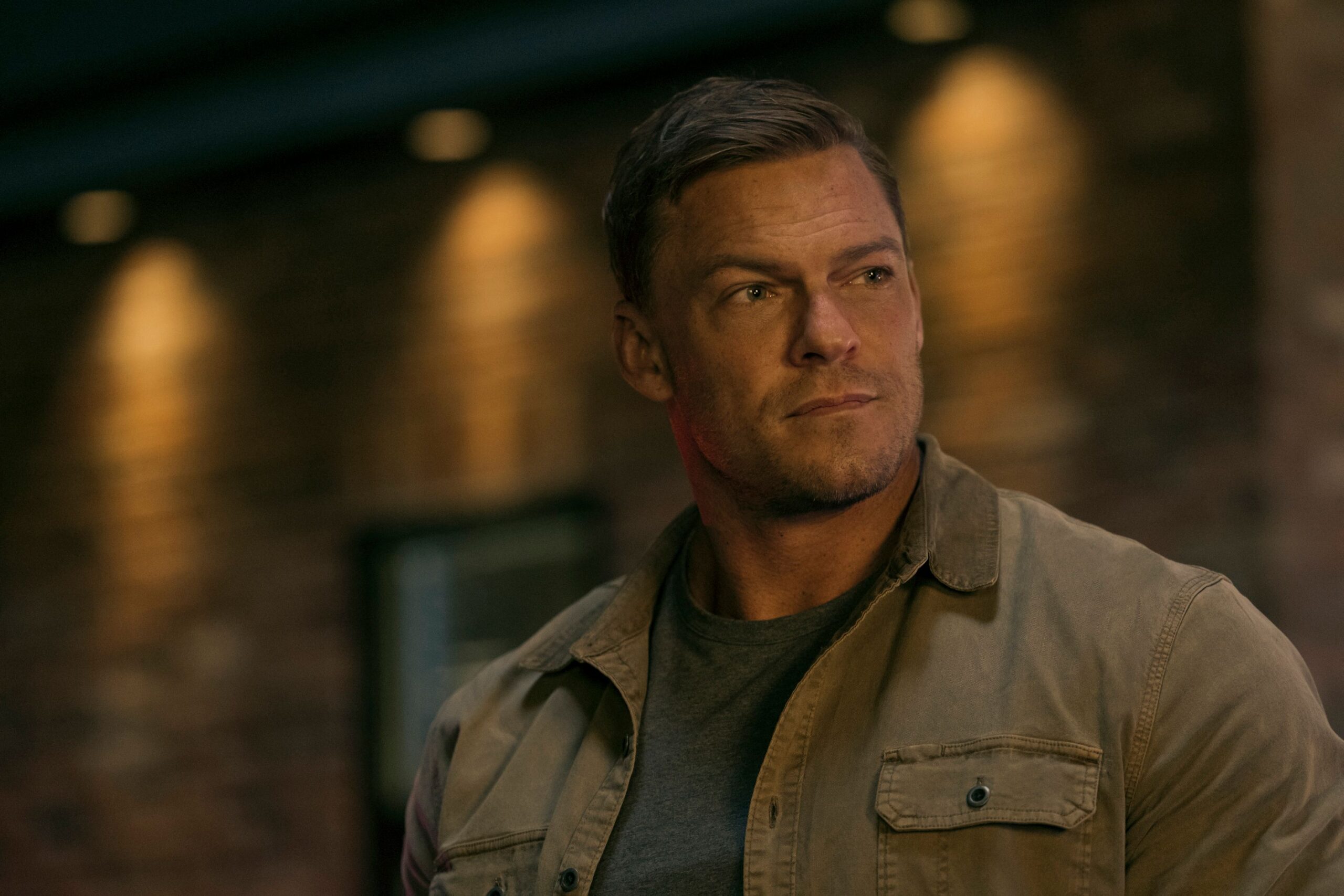A close up of Jack Reacher, a very large man, wearing a jacket and looking off in the distance, probably at someone smaller than he