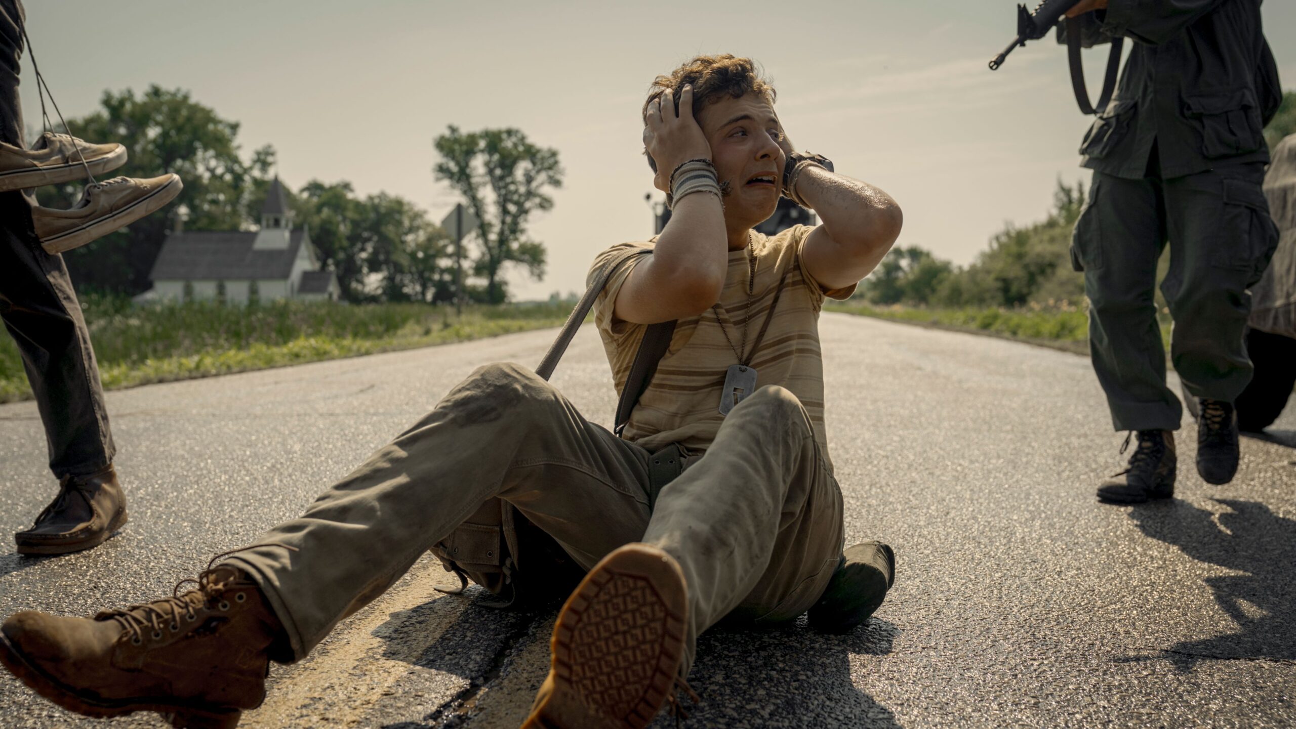 A skinny teenager sits in the road, clutching his head and crying, as soldiers carrying rifles approach him in The Long Walk