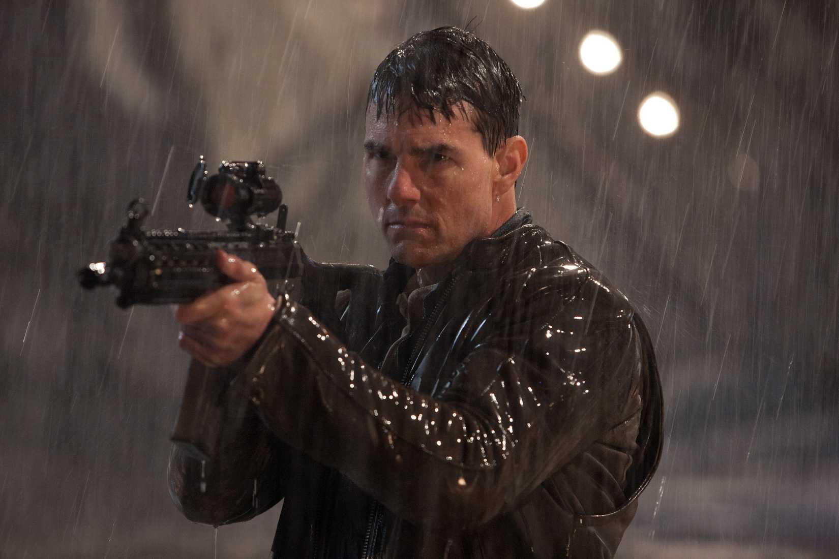 Tom Cruise stands in the rain holding a big gun