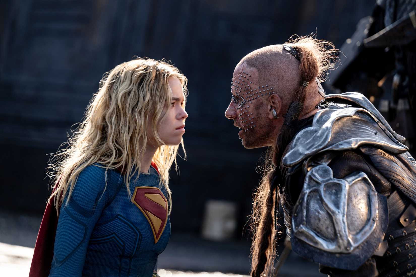 Milly Alcock as Supergirl, in the traditional costume, faces off against Krem, a man wearing patched-together metal armor, a shaved head and long topknot ponytail, and an extensive grid of facial piercings