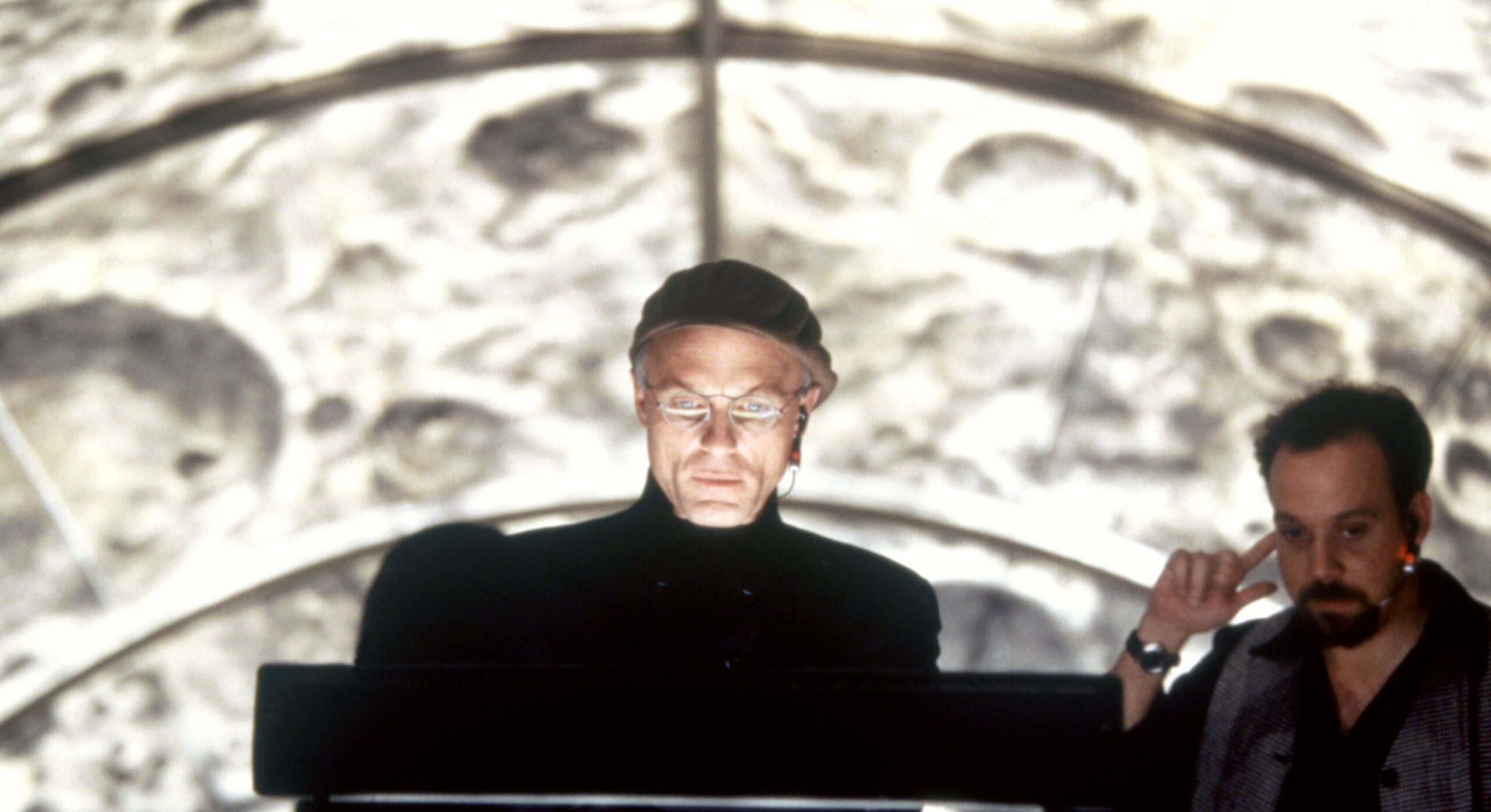 Ed Harris in a beret looks into a bright TV monitor; behind him are the craters of the moon
