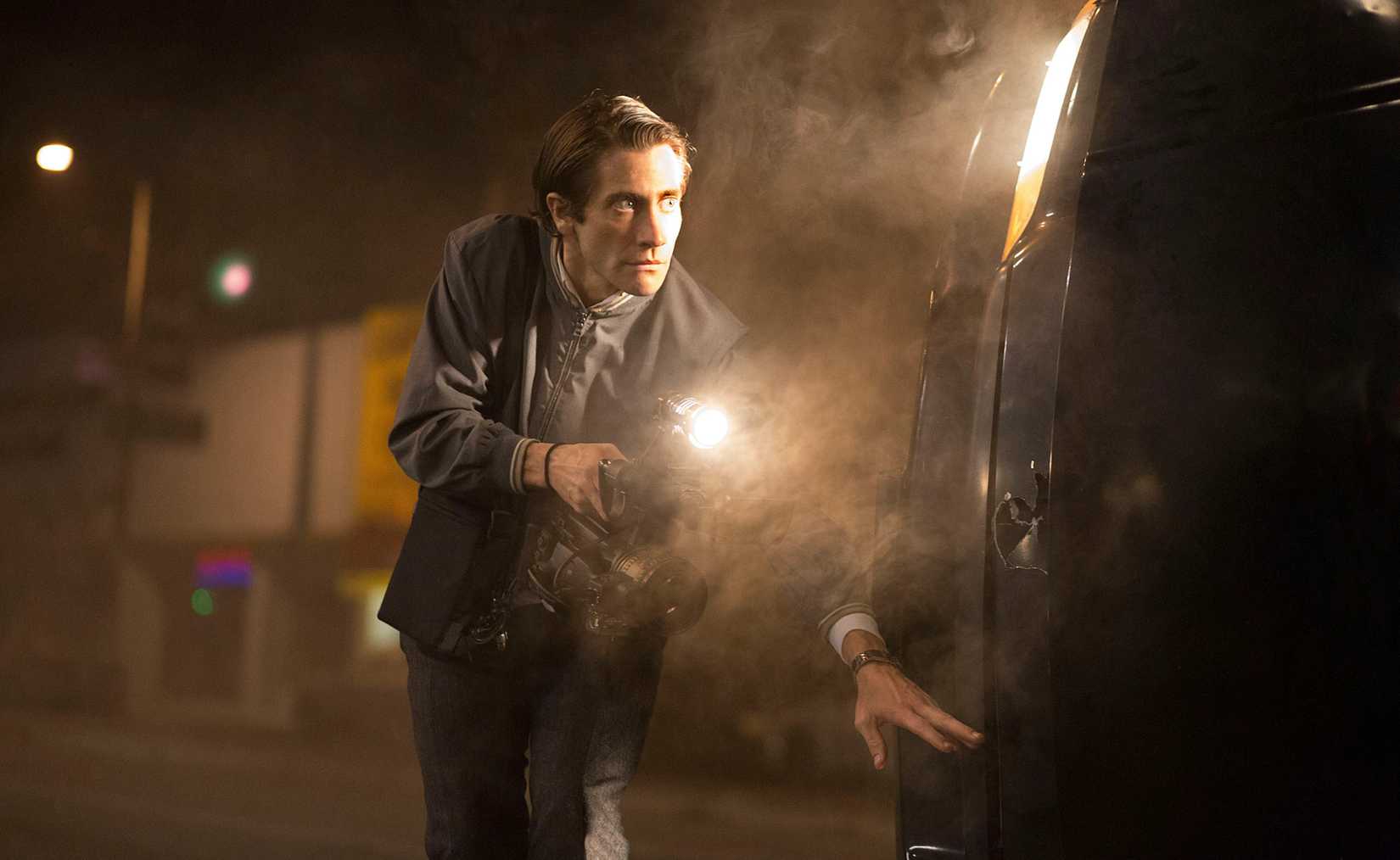 Jake Gyllenhaal as Louis “Lou” Bloom holding a camera behind an overturned car in Nightcrawler.