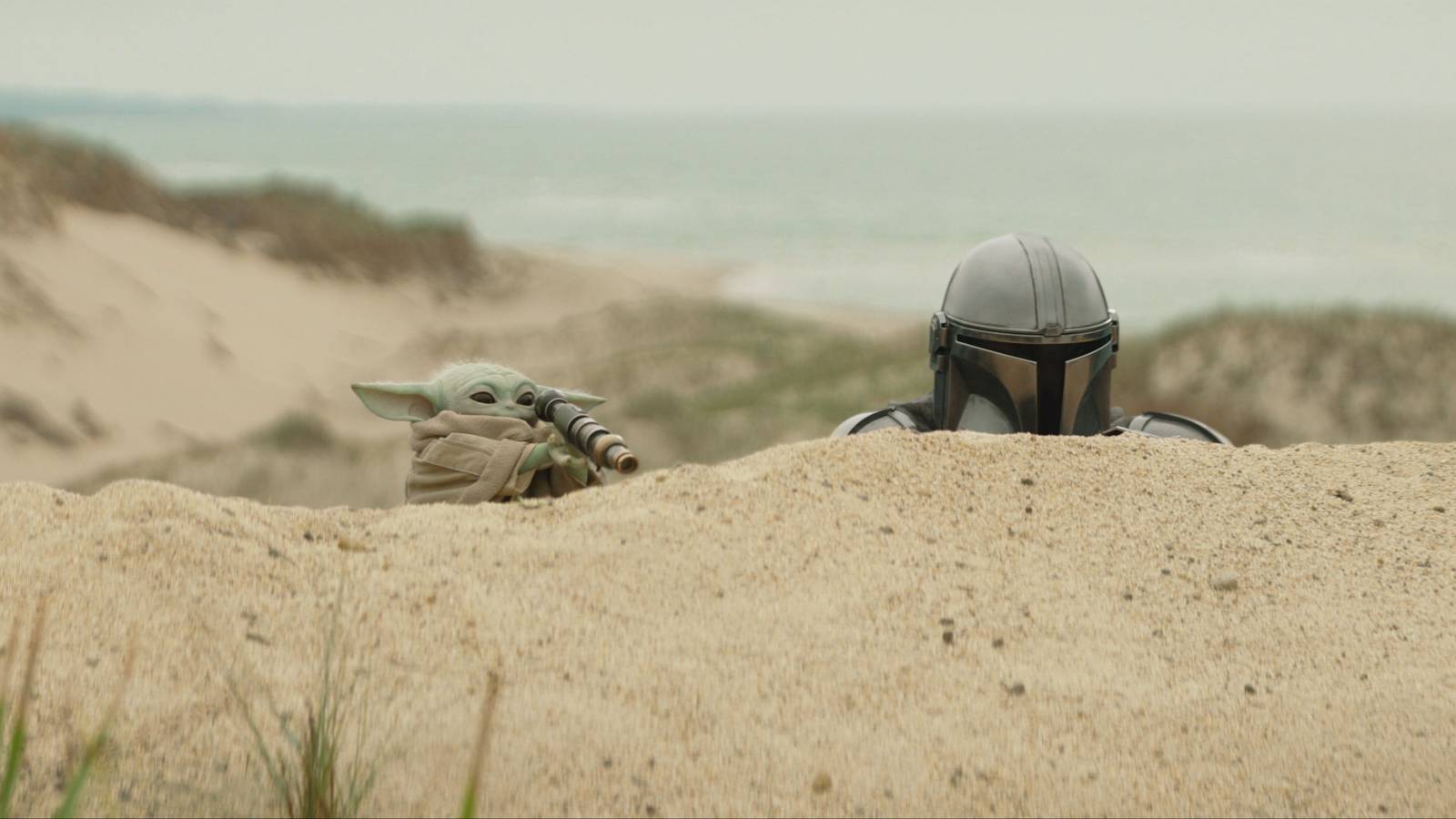 Grogu holds a spyglass while peeking over a sand dune alongside Din Djarin in The Mandalorian and Grogu.