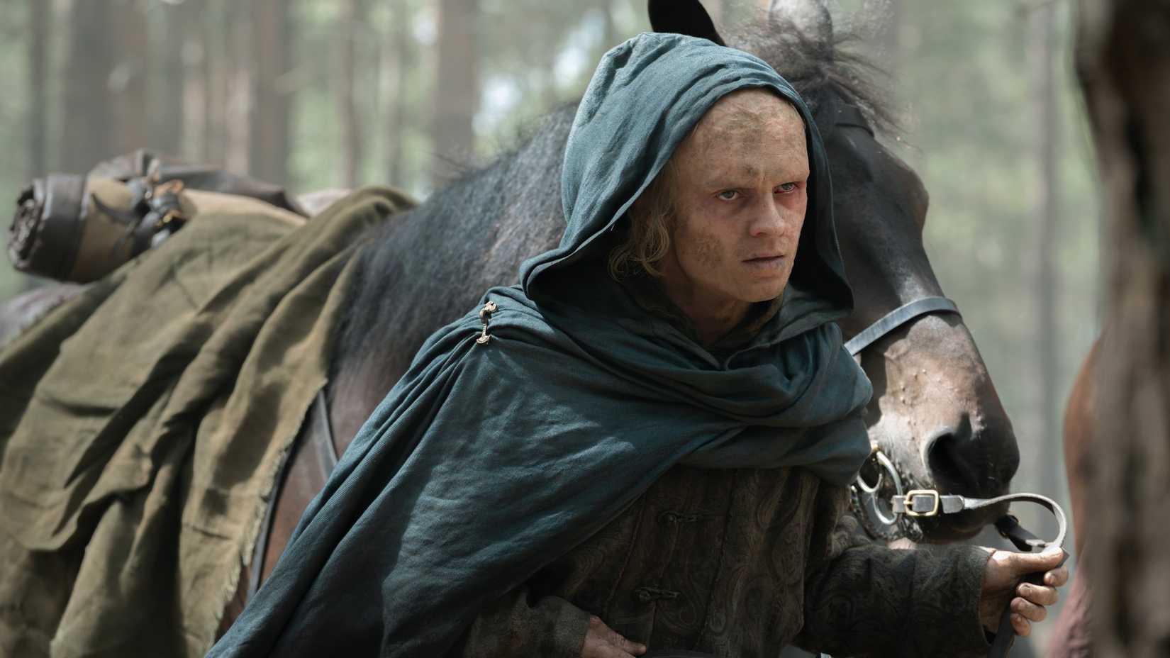 King Aemond (Tom Glynn-Carney) wearing a blue cloak leads a horse by its bridle in House of the Dragon season 3.