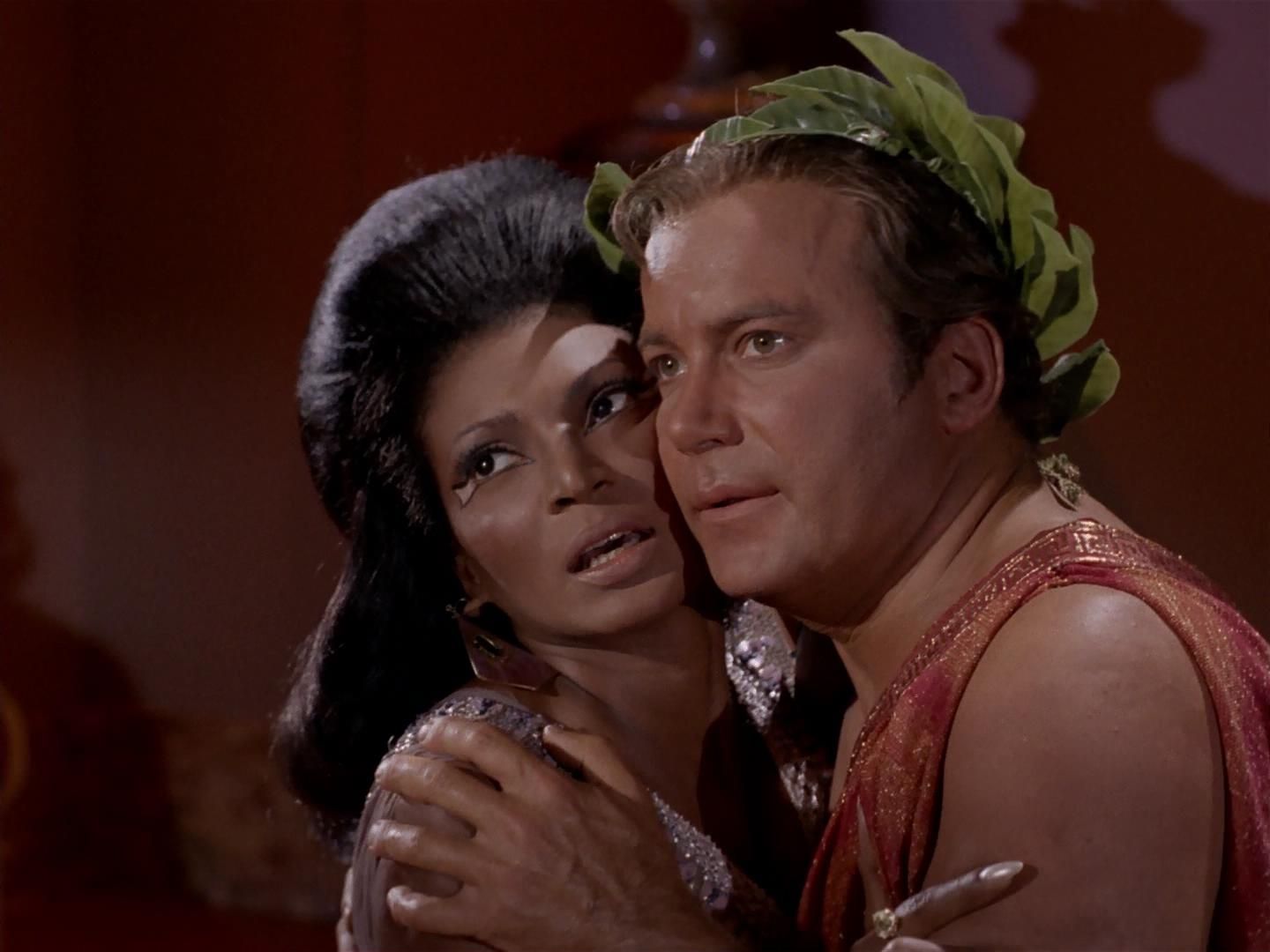 Uhura and Kirk look apprehensive and disturbed as the press their cheeks together and look off-camera. They are both dressed in a colorful imitation of ancient Roman dress, in Star Trek: The Original Series