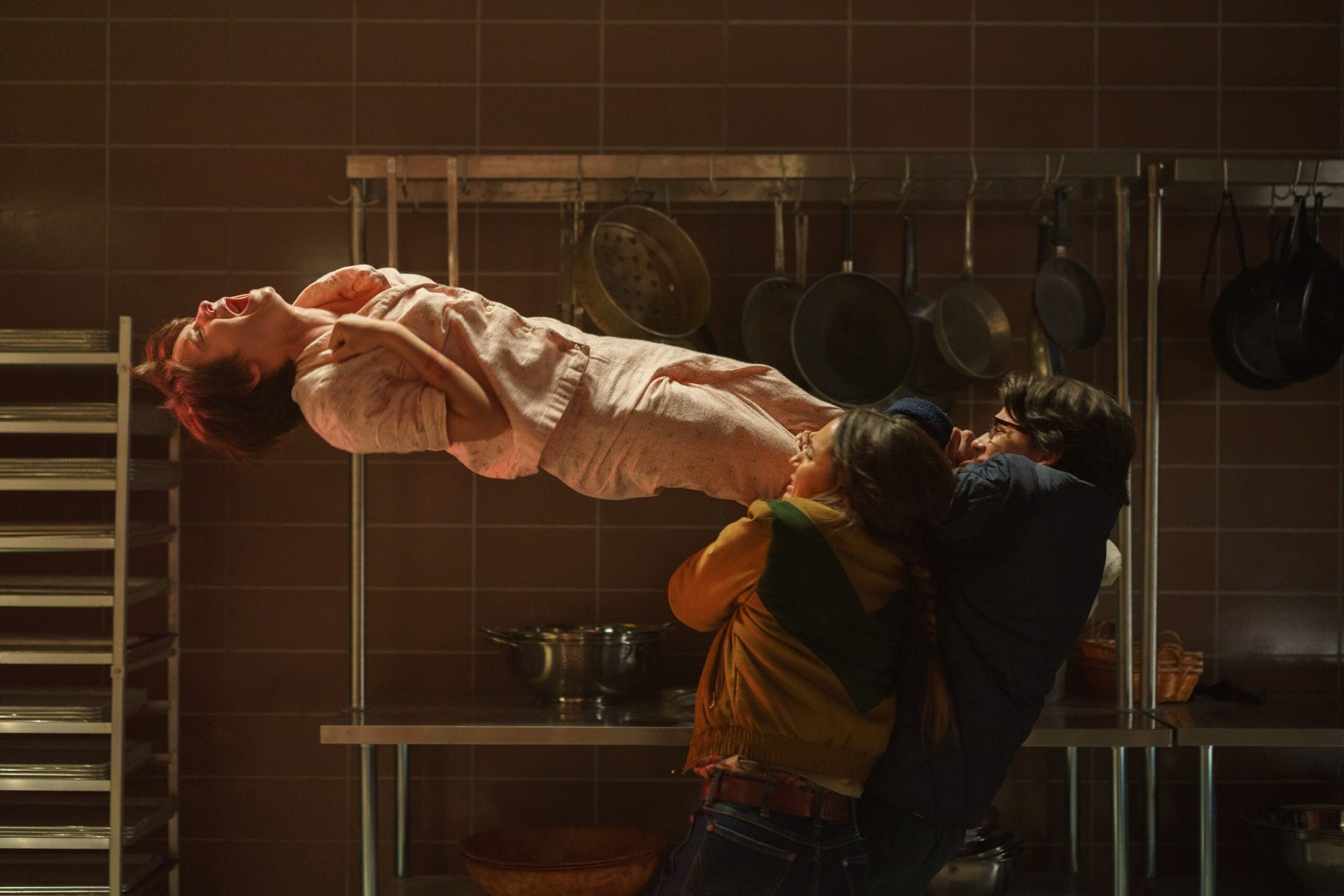 Gwen (Madeleine McGraw), in pink pajamas, floats on her back in the air, screaming and clutching at her throat, as several people cling to her legs and try to pull her down in Black Phone 2