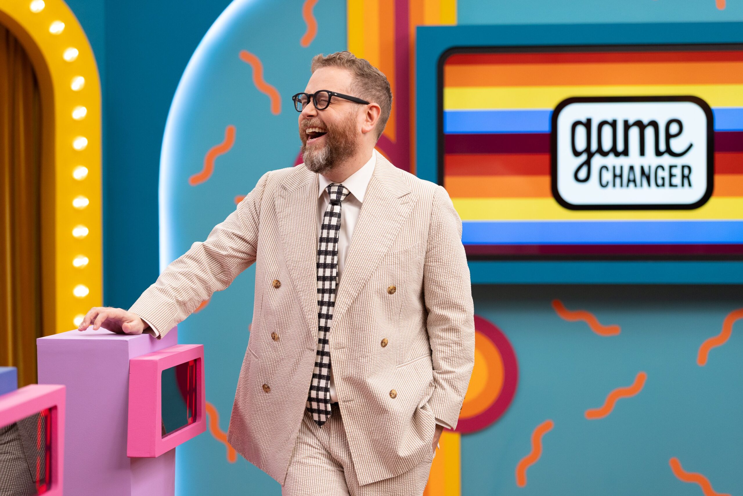 Dropout CEO and Game Changer host Sam Reich, wearing an off-white suit, laughs as he stands by a podium on the set of Dropout's comedy show Game Changer