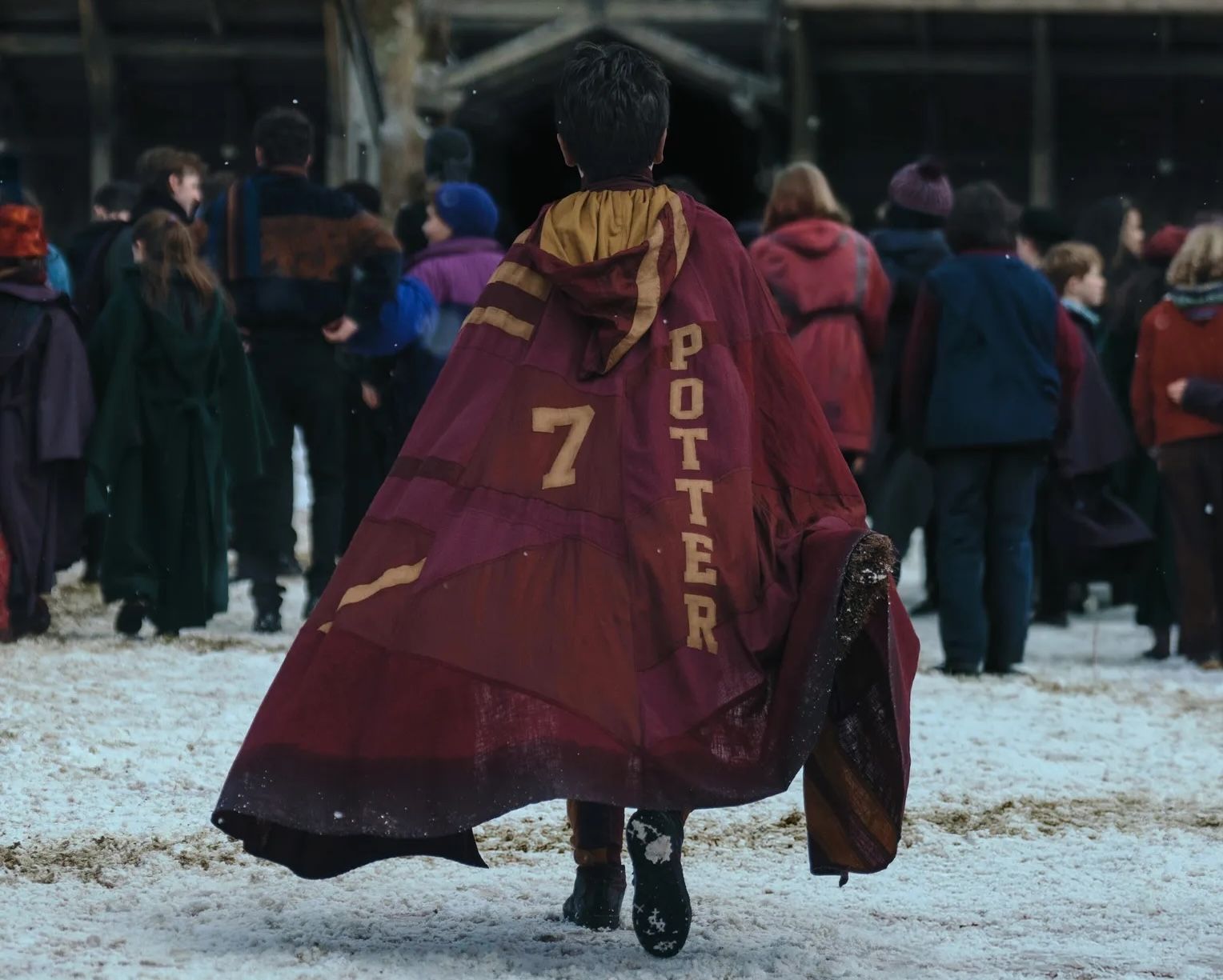 Harry Potter (Dominic McLaughlin), seen from behind as he walks across a snowy courtyard into Hogwarts, with a crowd of other out-of-focus students in the background. He's wearing a cloak that says "7" and "Potter" in the Gryffindor House colors.