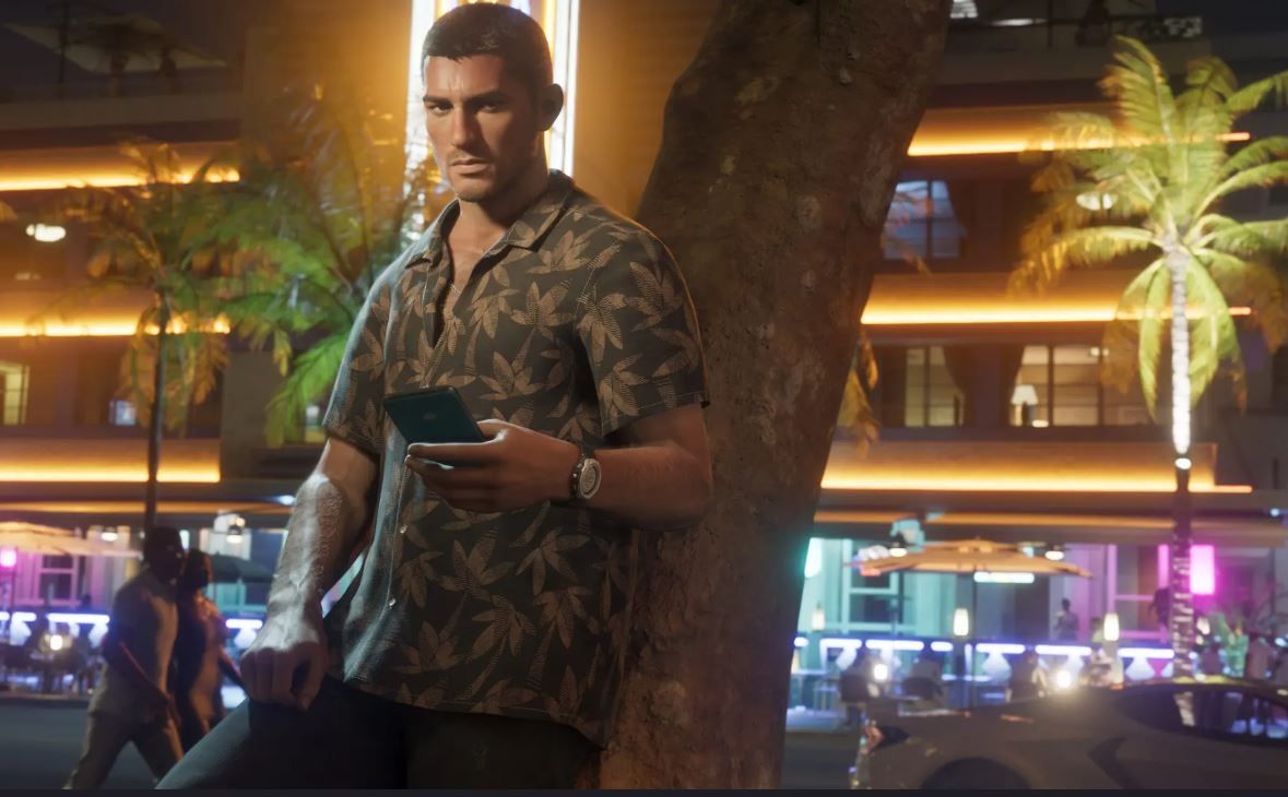 One of two protagonists in GTA 6, Jason, holds a phone as he leans against a tree. He's wearing a patterned shirt with leaves as the city nightlife bustles in the background.