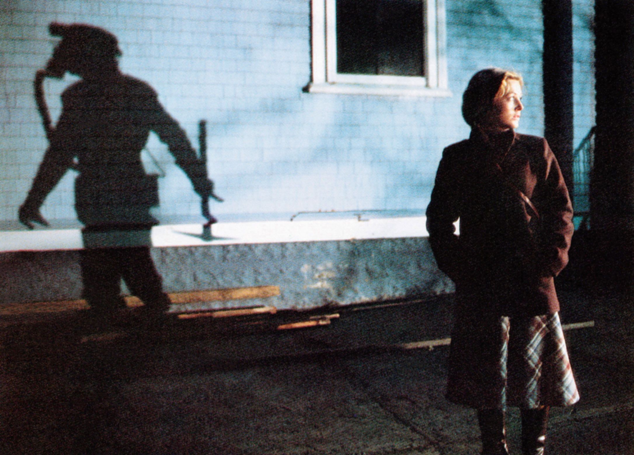 An image from the 1981 film My Bloody Valentine. It shows a blonde woman dressed in a brown jacket and plaid skirt looking behind her as a shadow of a miner with a pickaxe is highlighted on the wall next to her.