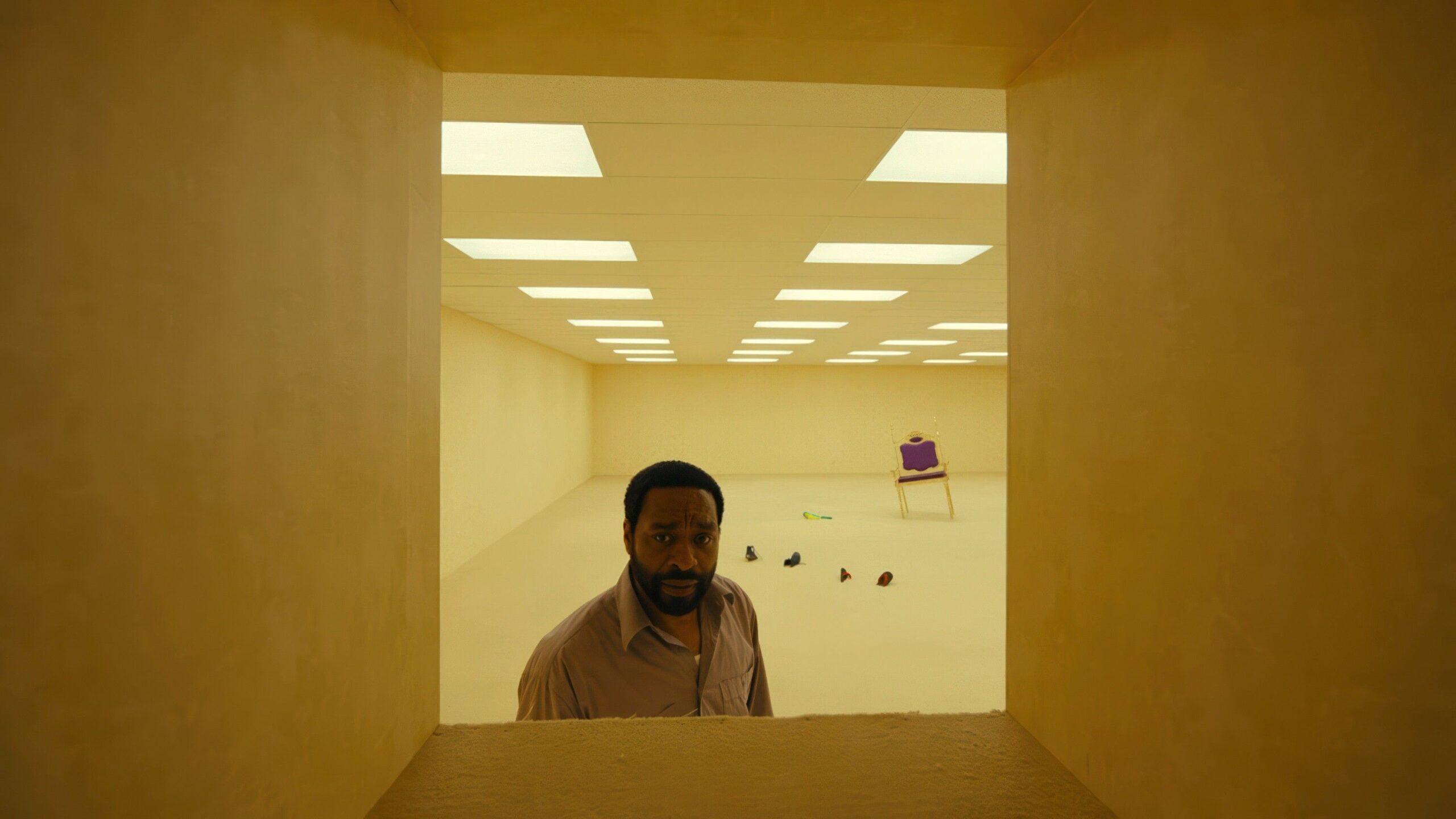 A Black man (Chiwetel Ejiofor) in a button-down shirt stands in a room with yellow wallpaper and a scattering of random objects on the floor behind him and peers through a chest-high, square opening in the wall in Backrooms