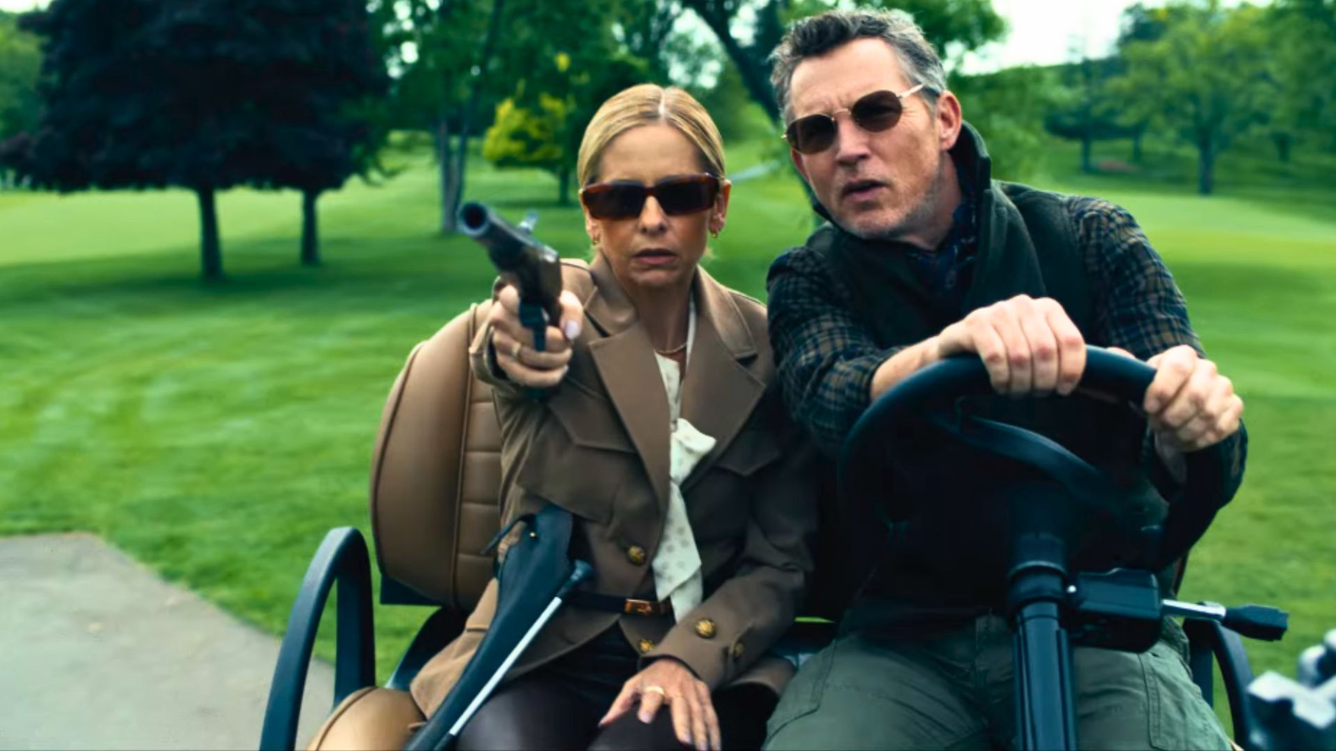 Sarah Michelle Geller, wearing sunglasses and a blazer, raises a gun to fire while sitting on what looks to be a golf buggy.