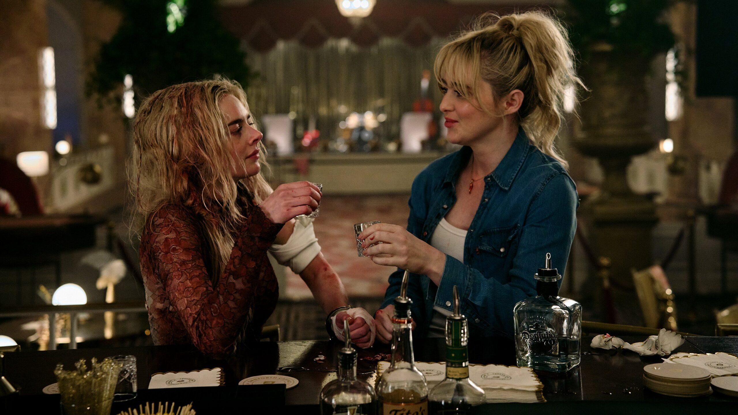 Grace (Samara Weaving), a woman wearing a bloadsoaked wedding dress, and Faith (Kathryn Newton) wearing a denim overshirt, share shots of tequila in a hotel ballroom