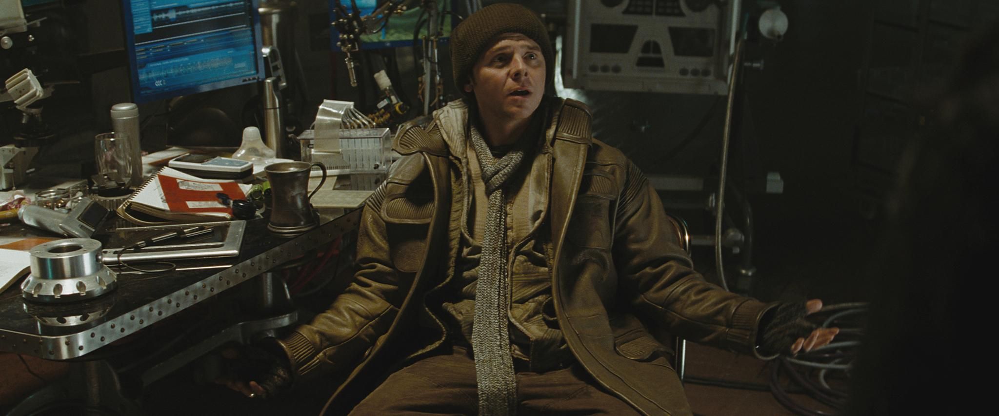 Simon Pegg as Scotty in Star Trek (2009) spreads his arms in a questioning gesture, sitting in warm clothing at a cluttered desk.