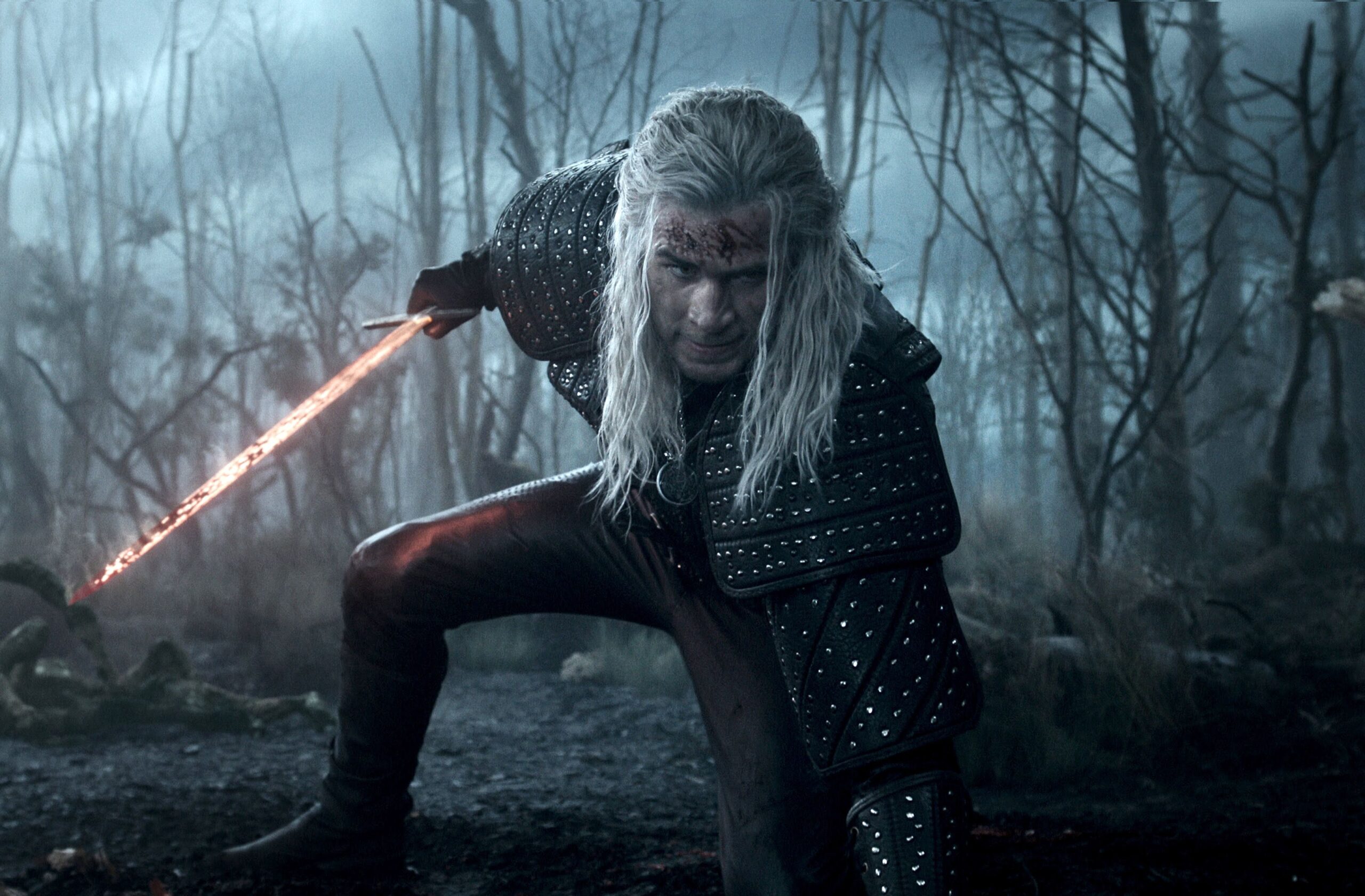 Liam Hemsworth as The Witcher crouching with a flaming sword in a blue-hued woods