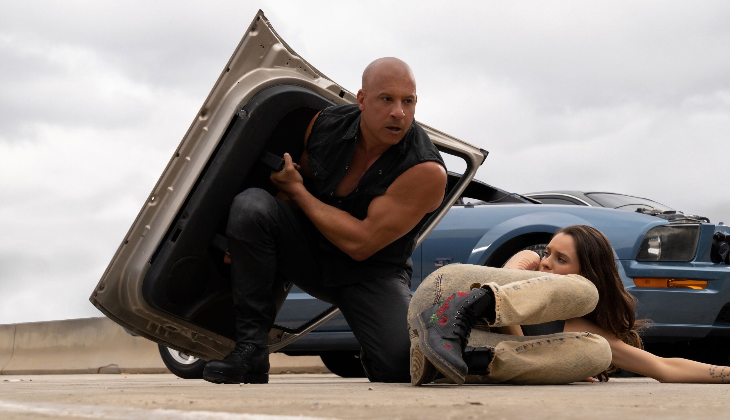 Dom Toretto (Vin Diesel) holds up a detached car door to shield himself from a hail of bullets as he moves to cover Isabel (Daniela Melchior), lying on the ground in front of a car, in Fast X