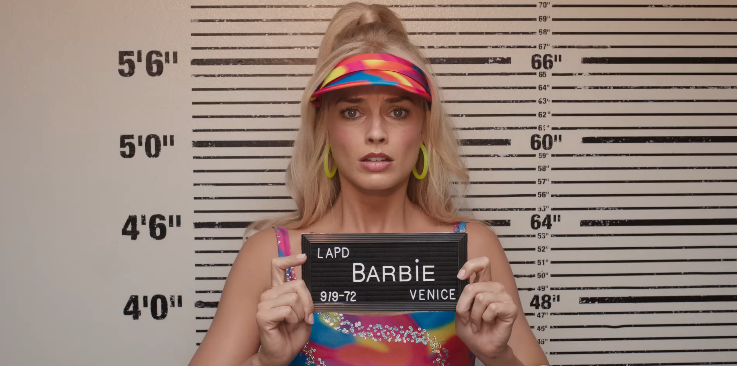 A still from the Barbie movie that shows Barbie taking a mugshot. Barbie is played by Margot Robbie and she looks distressed as she holds up a sign that says Barbie LAPD Venice.