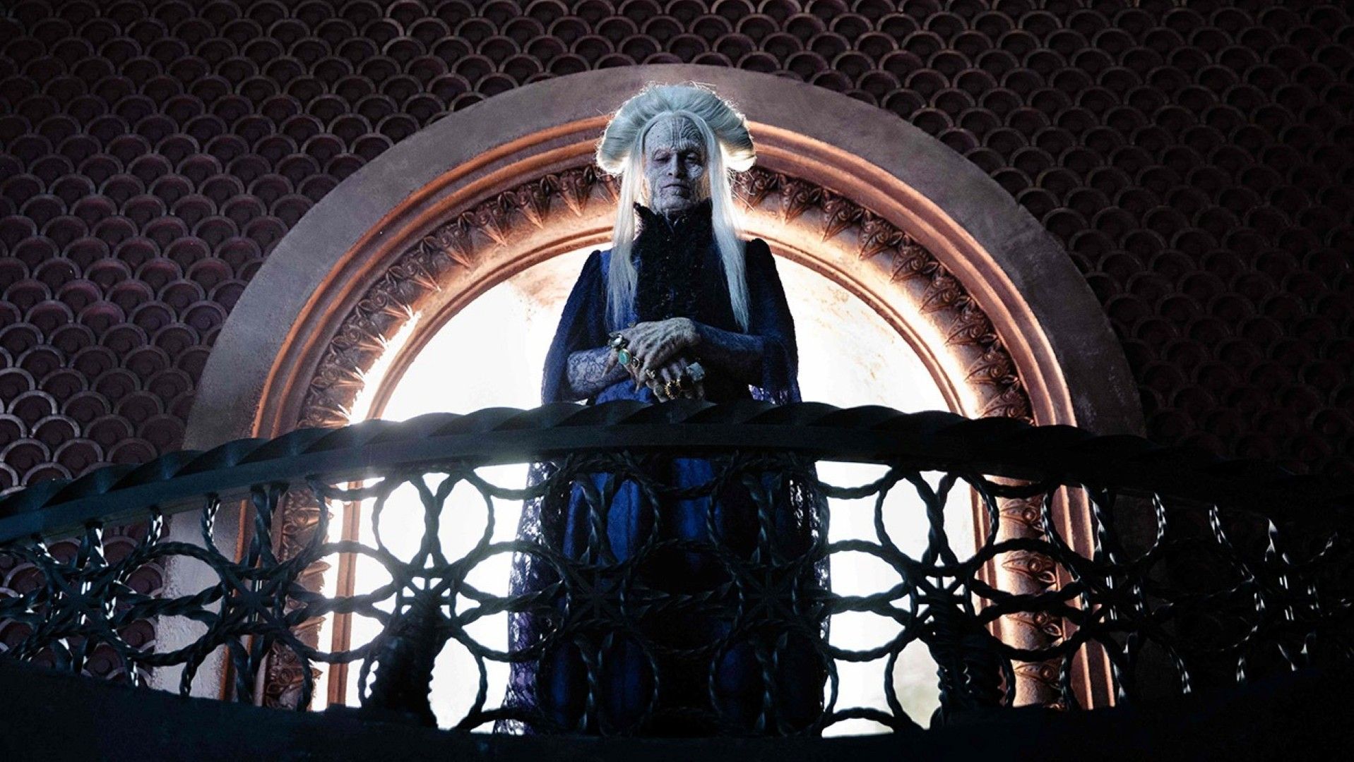 In a scene from Luc Besson's Dracula, Caleb Landry Jones, playing the title monster, stands on an ornate balcony in front of a window, wearing his white hair "up" in a way that looks like Gary Oldman's version of the character. 