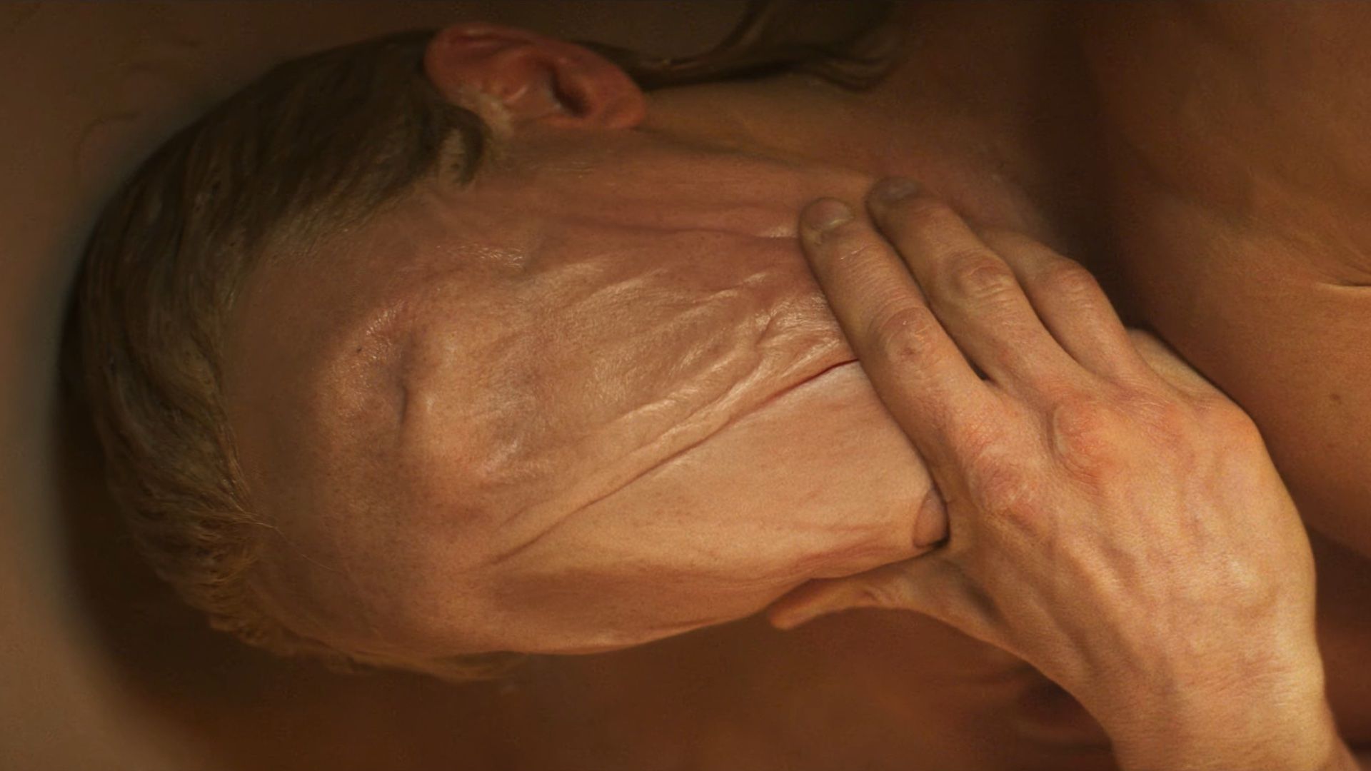 A closeup shot of Clayface, with his hand on his chin and an unbroken flap of skin from forehead to chin, from the movie's first teaser trailer