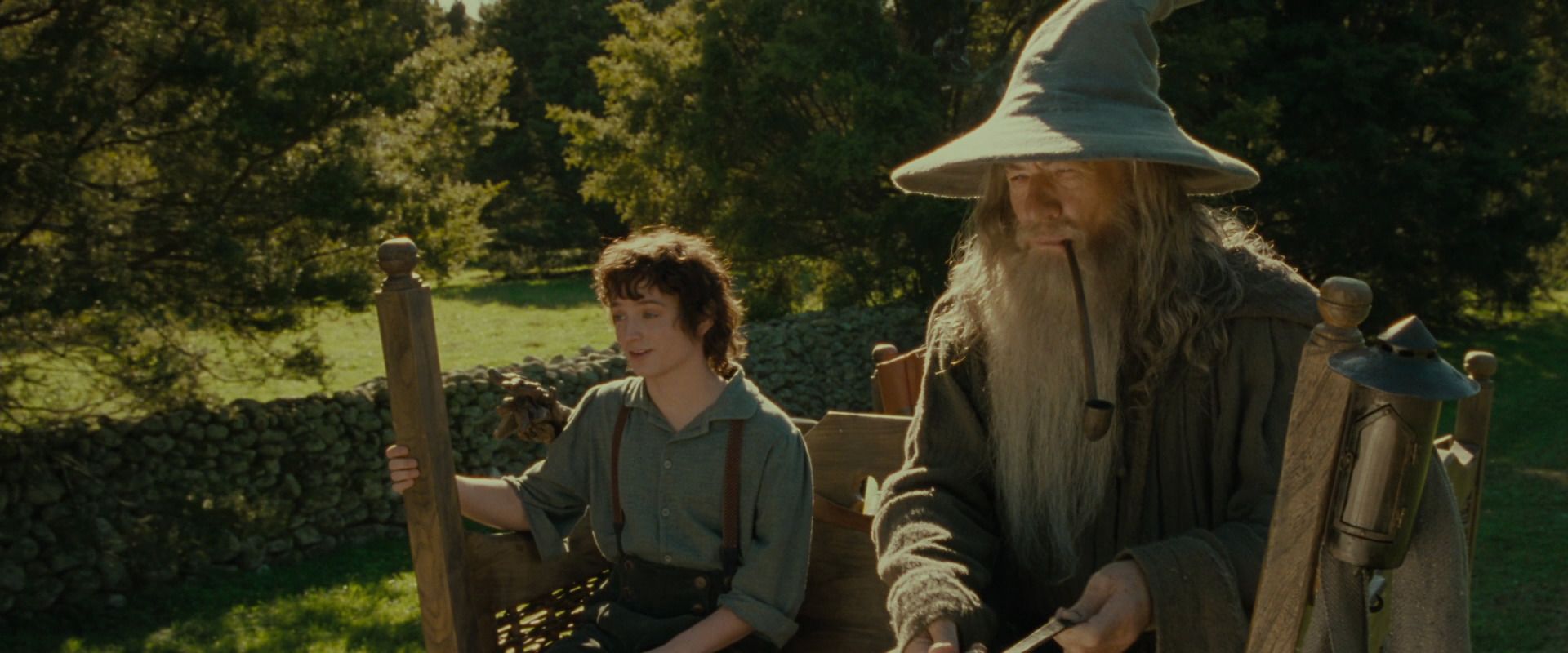 A short man (Elijah Wood) sits on a cart next to a tall wizard (Ian McKellen) in a gray hat holding the reins of a horse.