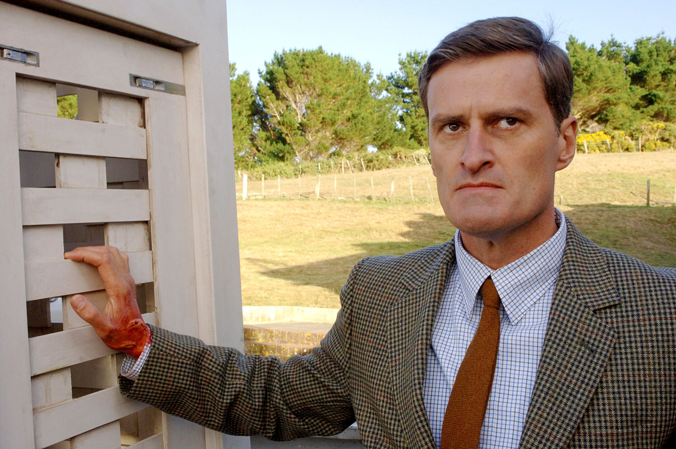 A man in a tweed suit looks ahead, annoyed.