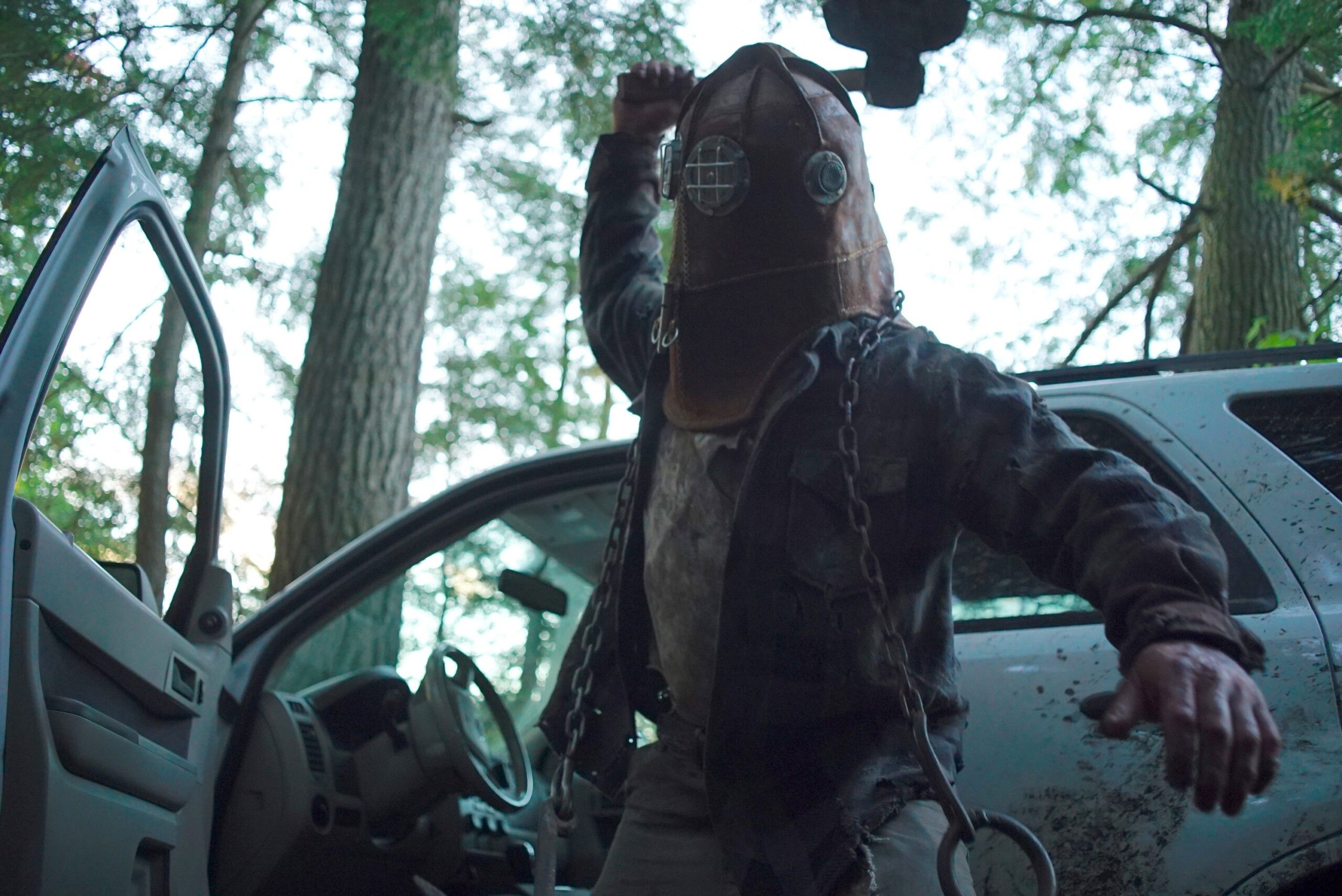 Johnny, a hulking man in a filthy leather jacket and diving-bell helmet, raises his axe to attack in In a Violent Nature