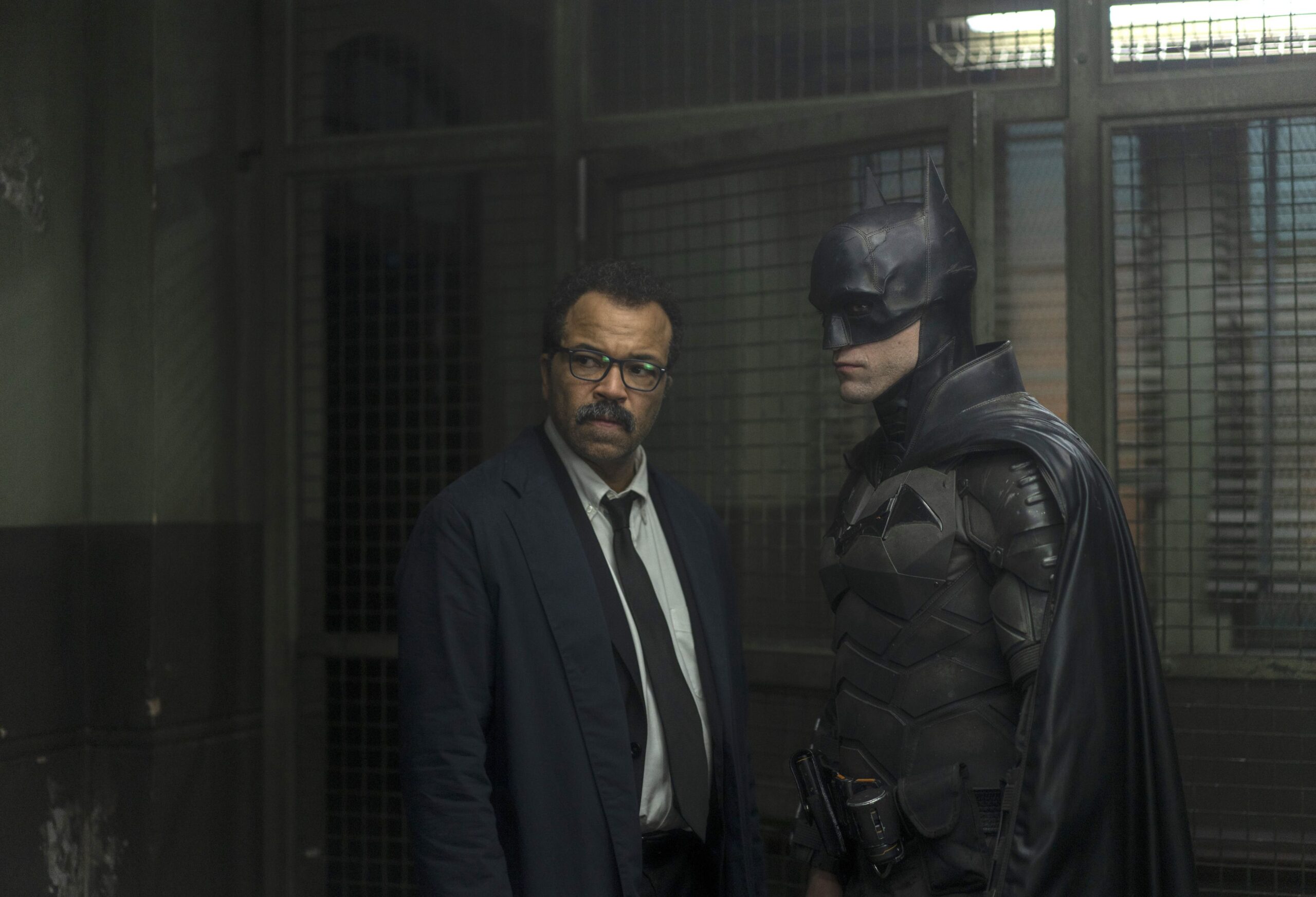 Left to right: Jeffrey Wright as Lt. James Gordon and Robert Pattinson as Batman in The Batman. They stand facing each other but faces turned out sternly inside a police precinct.