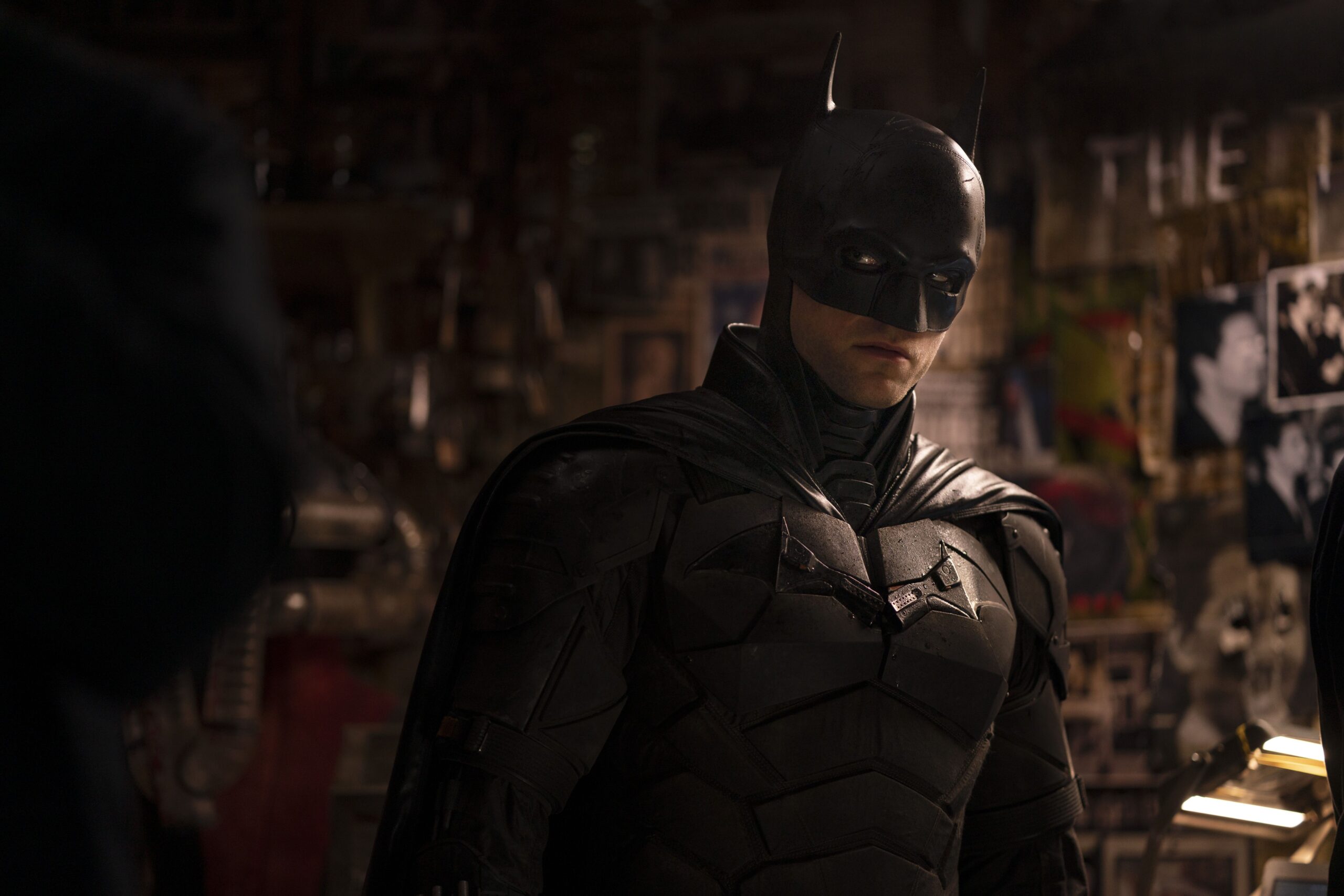Batman in black armor wearing a mask with batlike horns (Robert Pattinson) in front of a wall plastered with newspaper clippings and graffiti in The Batman