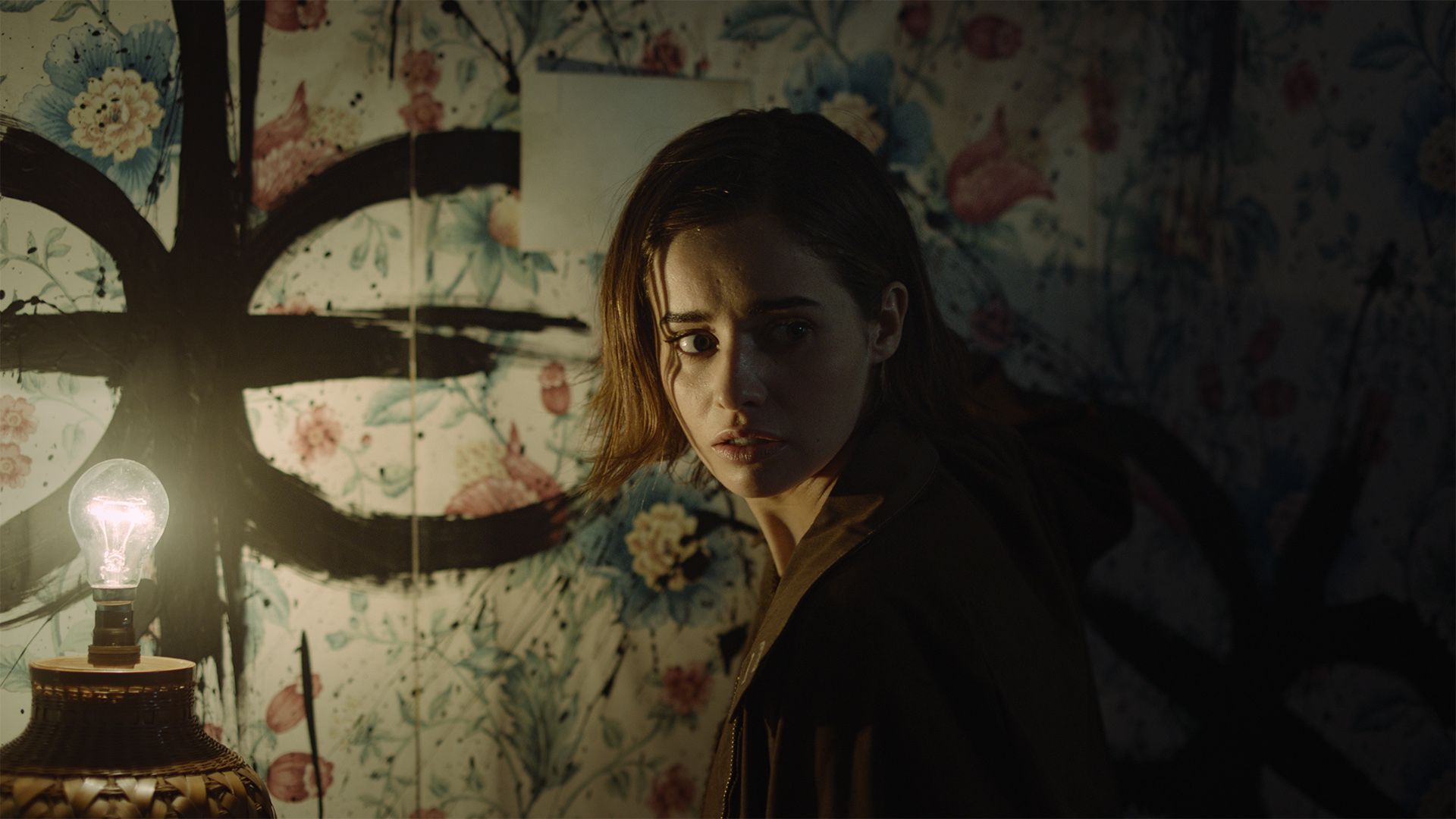 Erica, a white woman with dark hair, stands in a dimly lit room. She is looking over her shoulder with a concerned expression. A symbol has been painted on the wall.