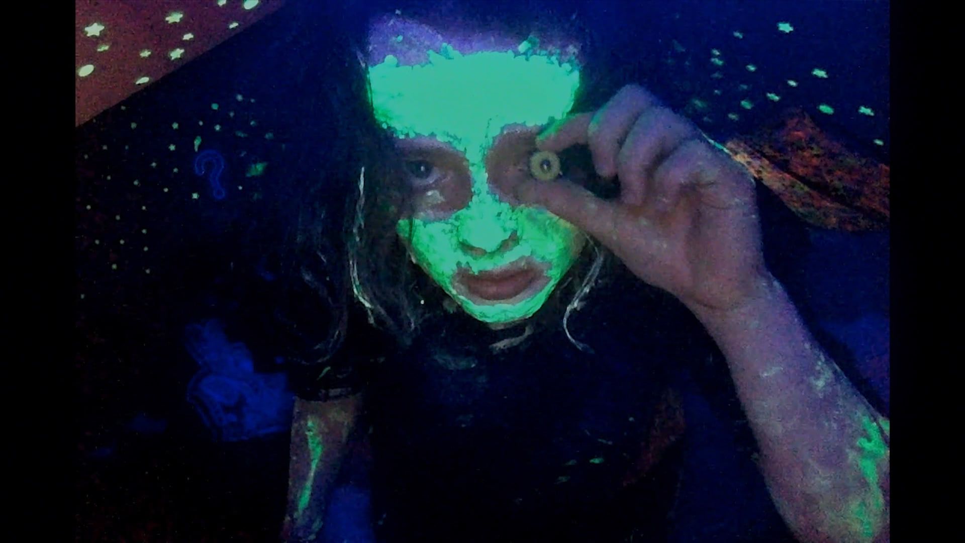Casey, covered in glow in the dark facepaint and holding a stuffed animal’s eyeball in front of her left eye, gazes ominously into her webcam in We’re All Going to the World’s Fair.